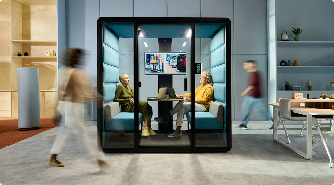 Two colleagues talking inside a soundproof meeting pod while coworkers walk past in an open-plan office