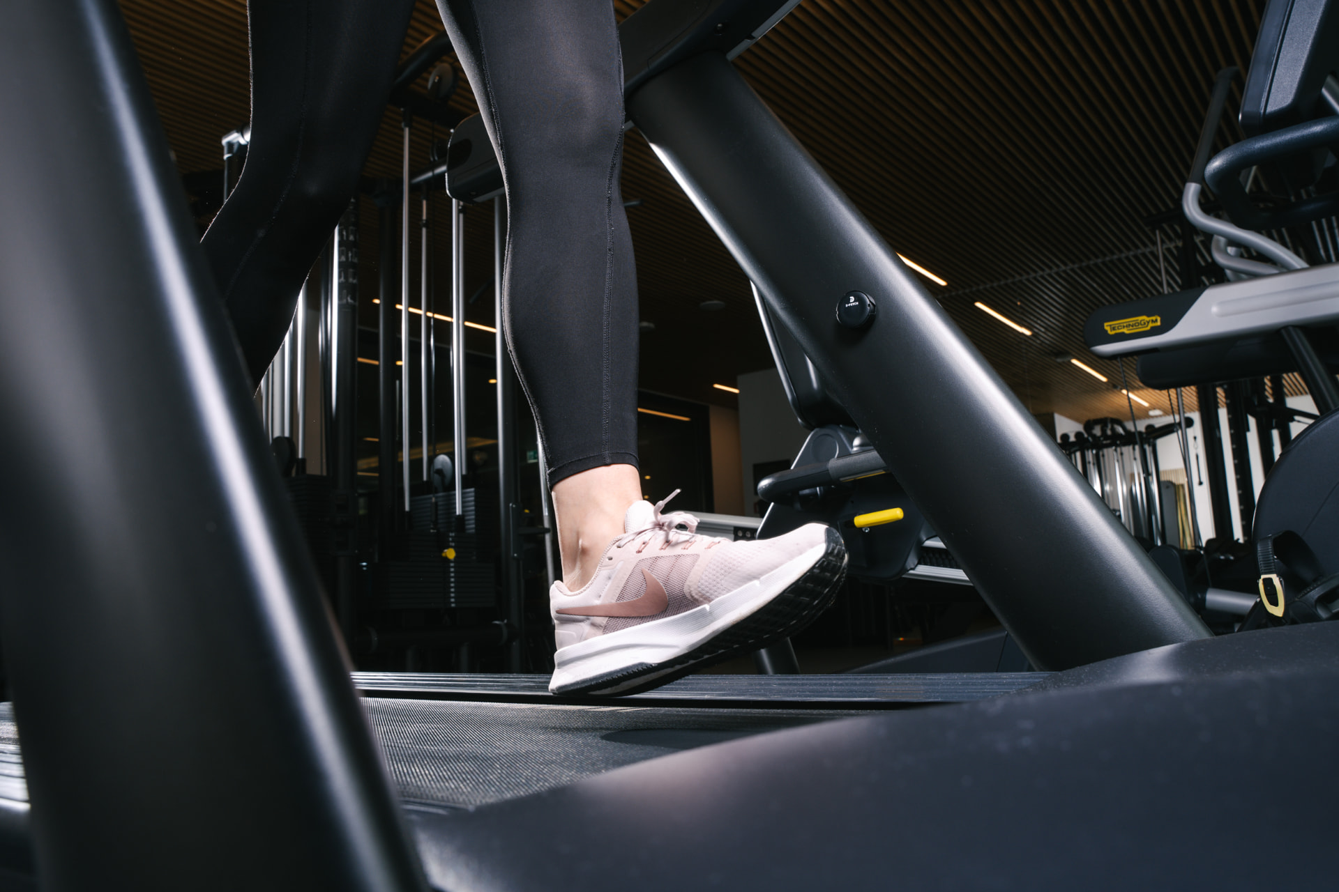 Low-angle view from a treadmill of a runner in black leggings and pink trainers, with weight machines and a Technogym cardio station behind